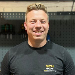mth-motoren-jost-hoeppener2 Jost Höppener - Junger Mann mit blonden hochgegelten Haare, schwarzem T-Shirt mit Firmenlogo steht vor Werkstatt